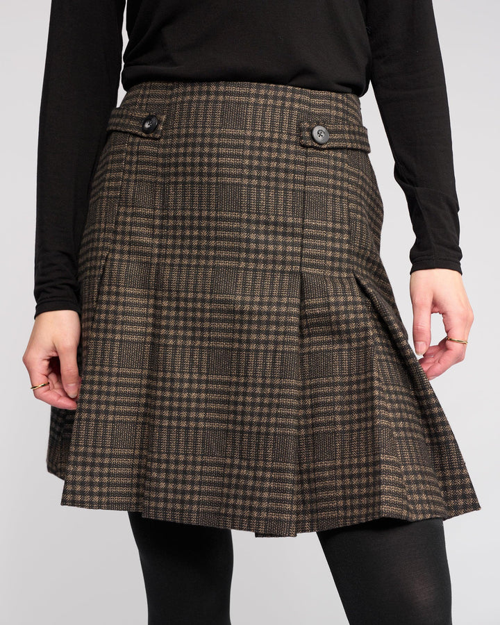 A model wears the Knewe Label Soho Skirt - Check, a brown and black checked pleated winter skirt with waist buttons, styled with a long-sleeve black top and black tights against a plain light background.