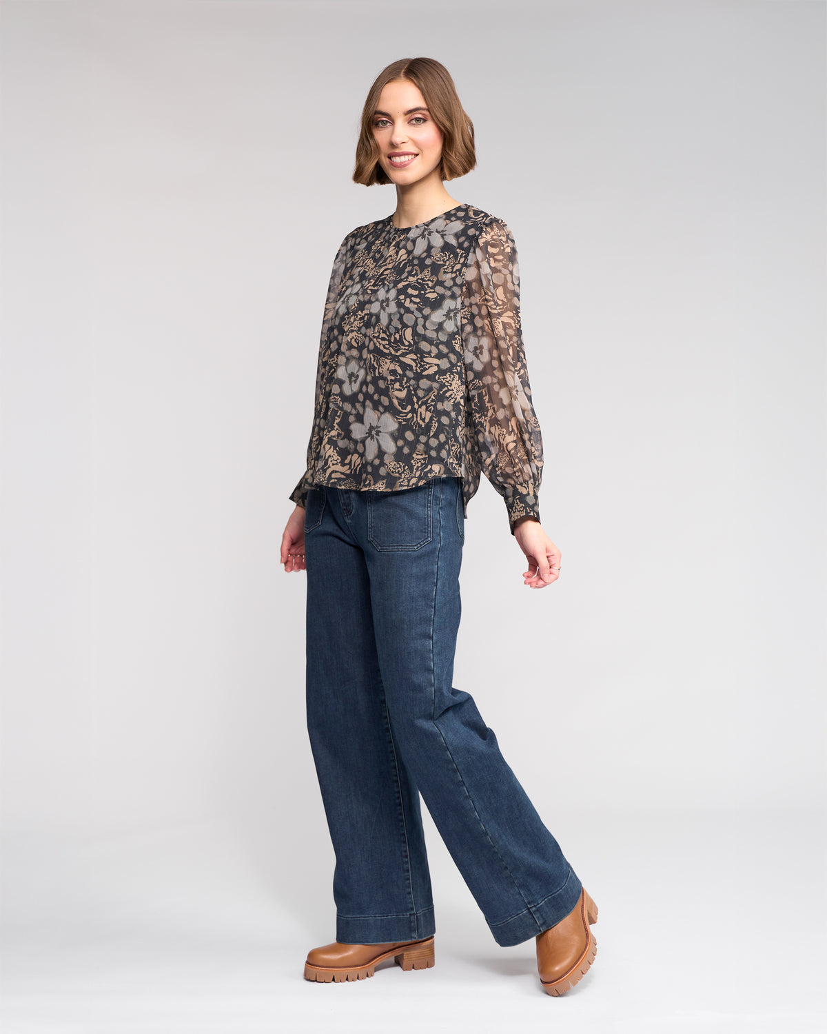 A woman smiles in the Lexie Top - Hazy by Knewe Label, a sheer long sleeve floral top, paired with wide-leg blue jeans and tan platform shoes against a light gray background.