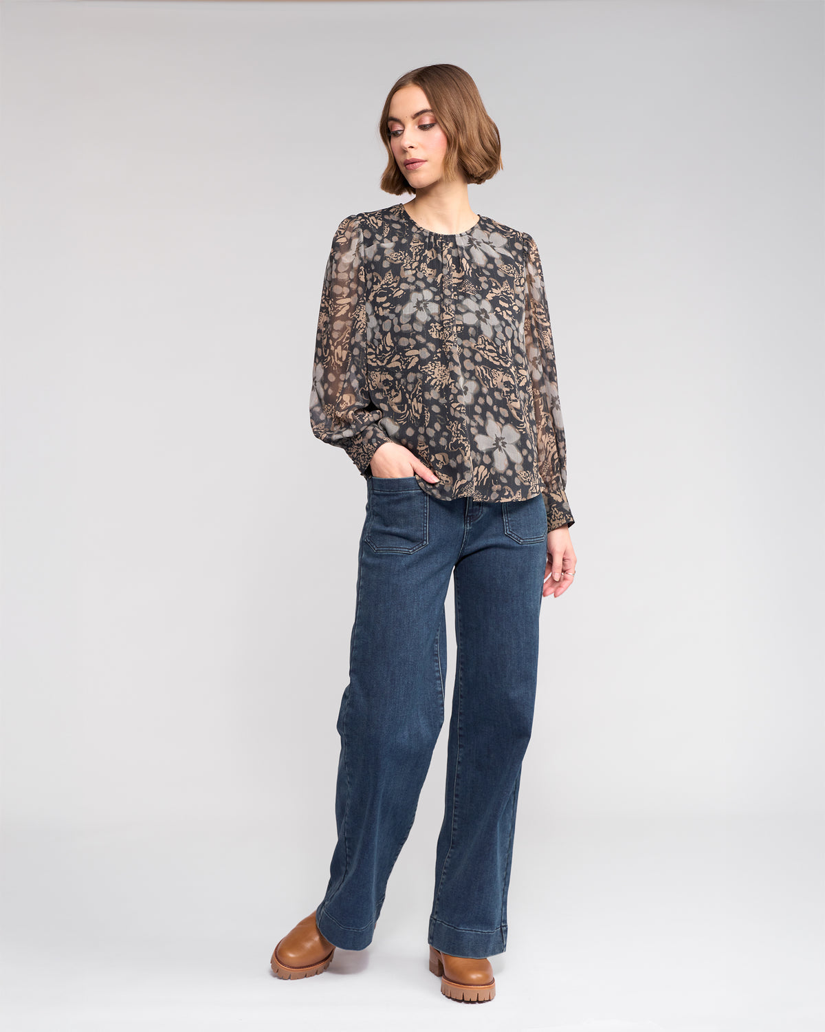 A woman with short brown hair stands against a plain background, wearing the Lexie Top - Hazy by Knewe Label, wide-leg blue jeans, and brown platform shoes. She has one hand in her pocket and looks slightly to the side.