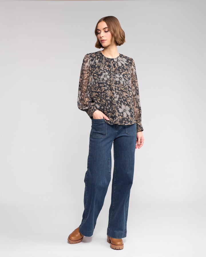 A woman with short brown hair stands against a plain background, wearing the Lexie Top - Hazy by Knewe Label, wide-leg blue jeans, and brown platform shoes. She has one hand in her pocket and looks slightly to the side.