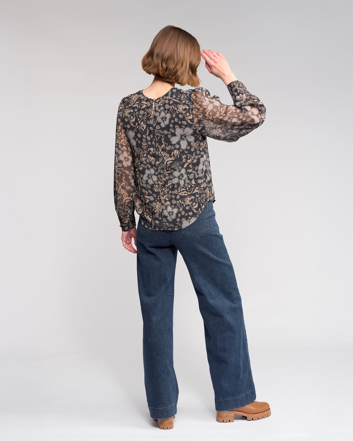 A person with short brown hair, wearing the Knewe Label Lexie Top - Hazy, blue jeans, and tan platform shoes, stands facing away with their right hand raised toward their head against a plain light gray background.