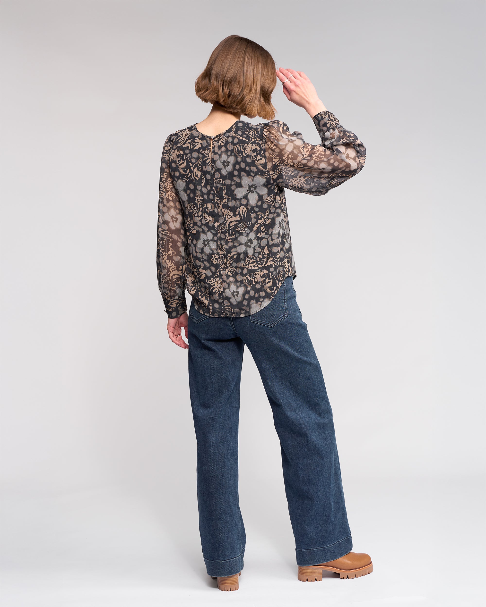 A person with short brown hair, wearing the Knewe Label Lexie Top - Hazy, blue jeans, and tan platform shoes, stands facing away with their right hand raised toward their head against a plain light gray background.