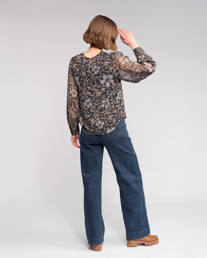 A person with short brown hair, wearing the Knewe Label Lexie Top - Hazy, blue jeans, and tan platform shoes, stands facing away with their right hand raised toward their head against a plain light gray background.