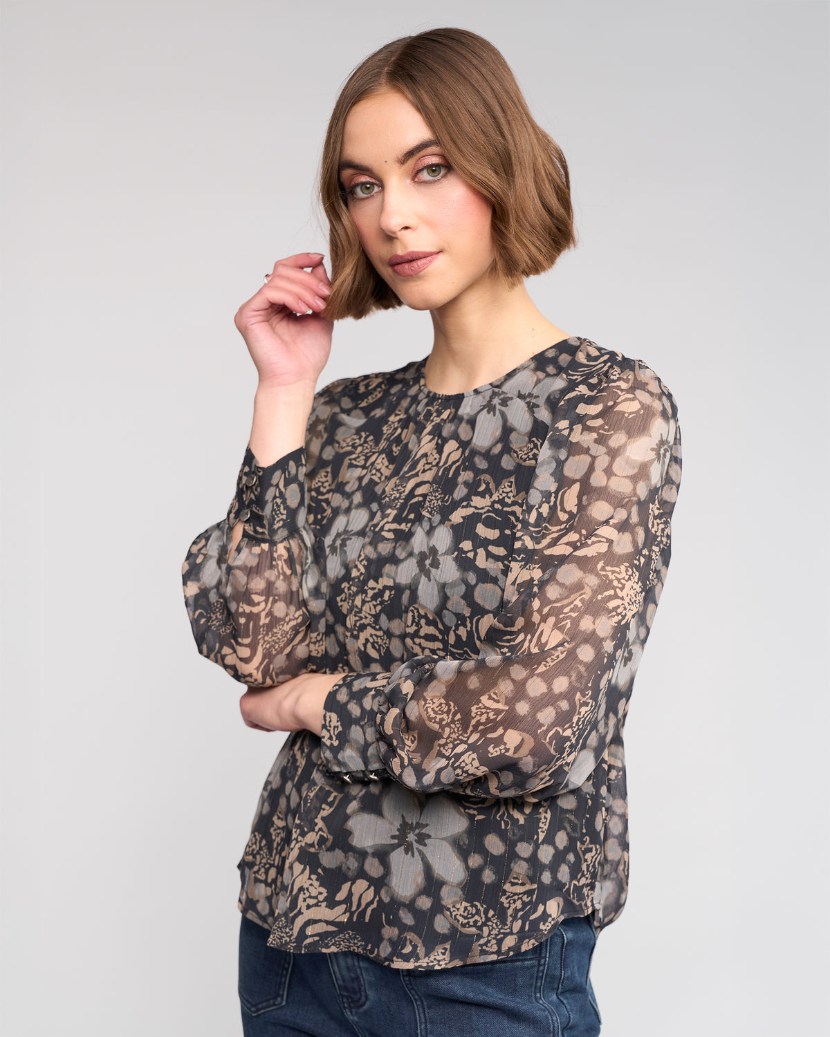 A woman with a short brown bob wears the Knewe Label Lexie Top - Hazy, a sheer long sleeve floral print blouse, paired with blue jeans. She stands against a plain light background, looking at the camera with a relaxed expression.