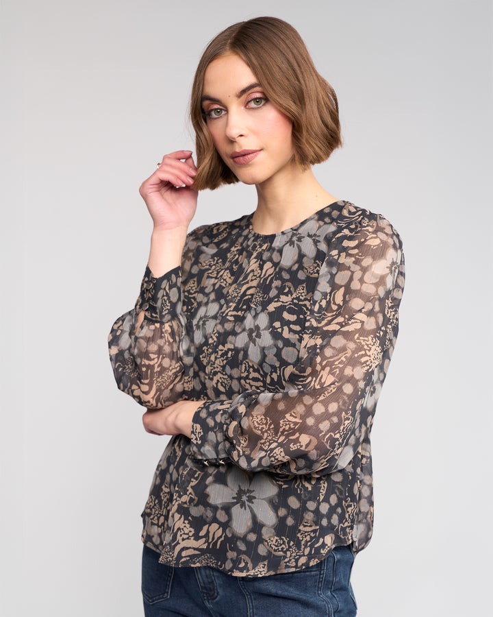 A woman with a short brown bob wears the Knewe Label Lexie Top - Hazy, a sheer long sleeve floral print blouse, paired with blue jeans. She stands against a plain light background, looking at the camera with a relaxed expression.