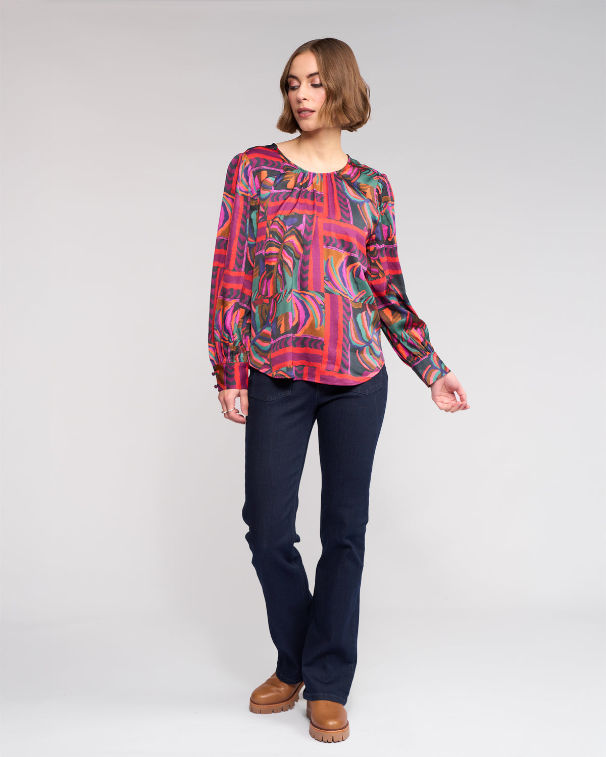 A woman stands against a plain background wearing the Knewe Label Lexie Top - Sunset, a colorful, sunset-print long sleeve in soft viscose-rayon blend, styled with dark jeans and brown platform shoes. She looks slightly to the side with one hand raised.