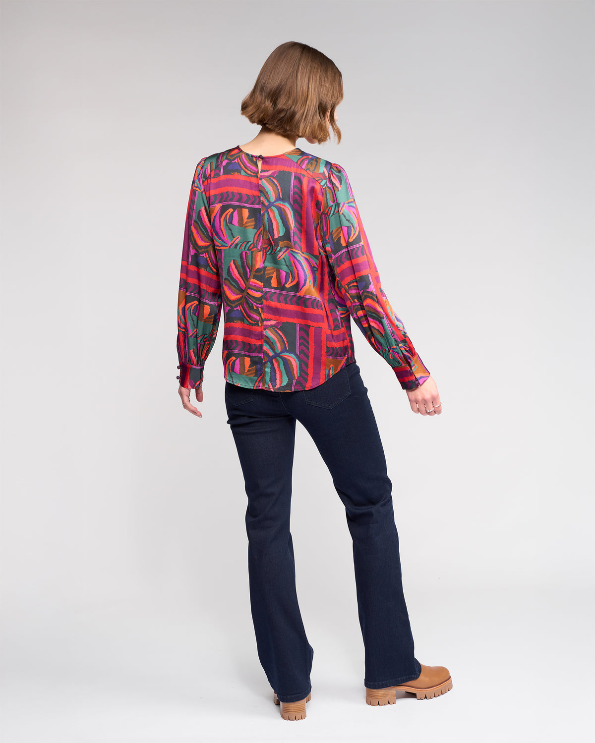 A person with chin-length brown hair stands facing away, wearing the Knewe Label Lexie Top in the Sunset print, paired with dark blue jeans and tan platform shoes, against a plain light background.
