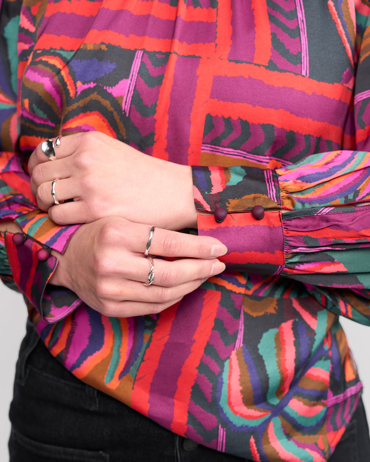 A person wearing the Knewe Label Lexie Top - Sunset, featuring a colorful abstract pattern and long sleeves, adjusts the cuff. They have several silver rings and dark pants. The background is plain and light-colored.