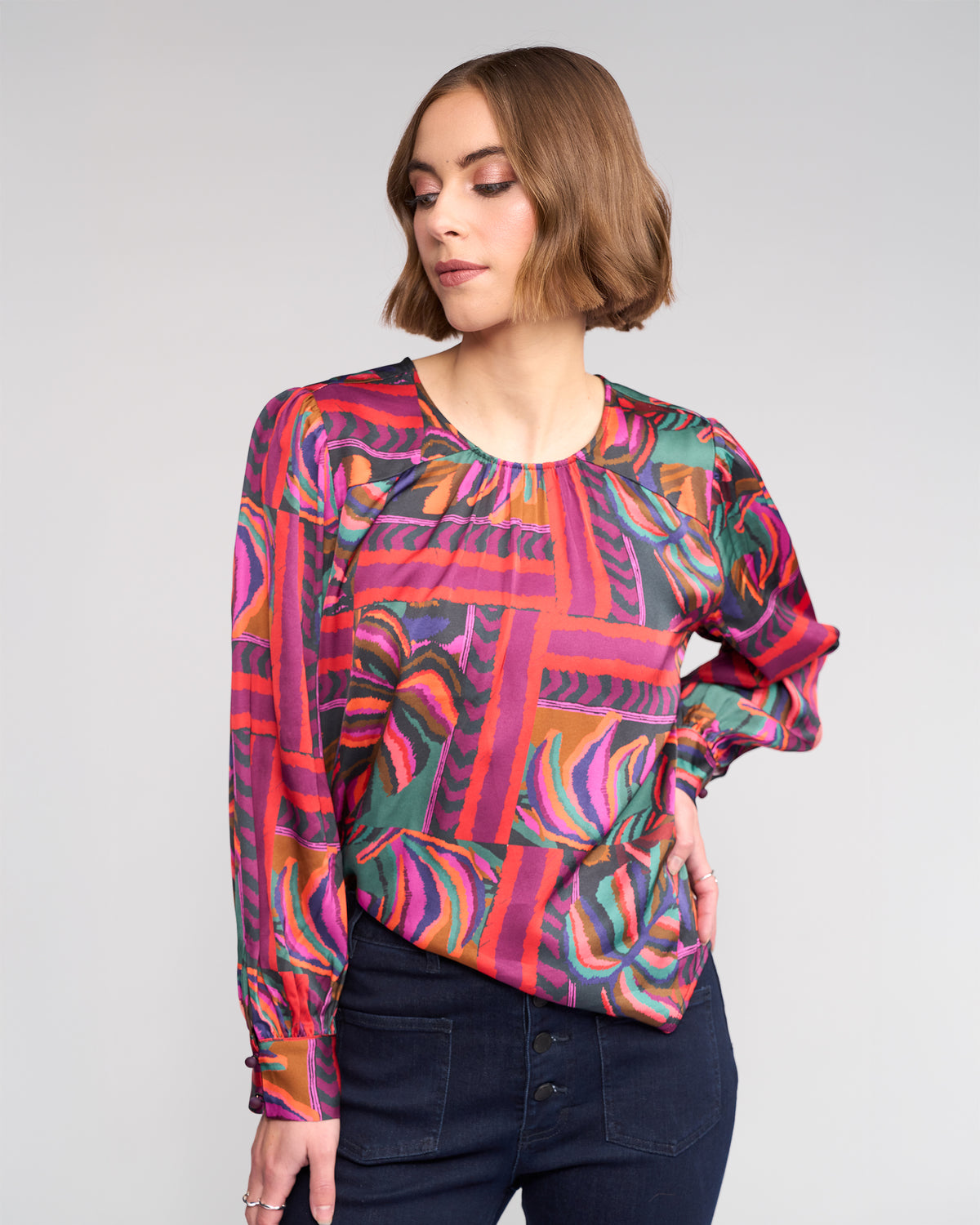 A woman with short brown hair models the Knewe Label Lexie Top - Sunset, a vibrant long-sleeve geometric print in pink, red, purple, and green. She pairs it with dark jeans and poses with one hand on her hip against a light gray background.