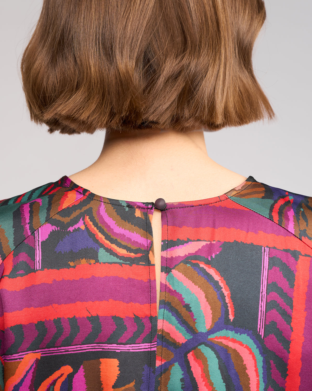 A close-up of the back of a person with short brown hair wearing the Knewe Label Lexie Top - Sunset, a colorful patterned long sleeve top with a keyhole button neckline, crafted from a soft viscose-rayon blend.