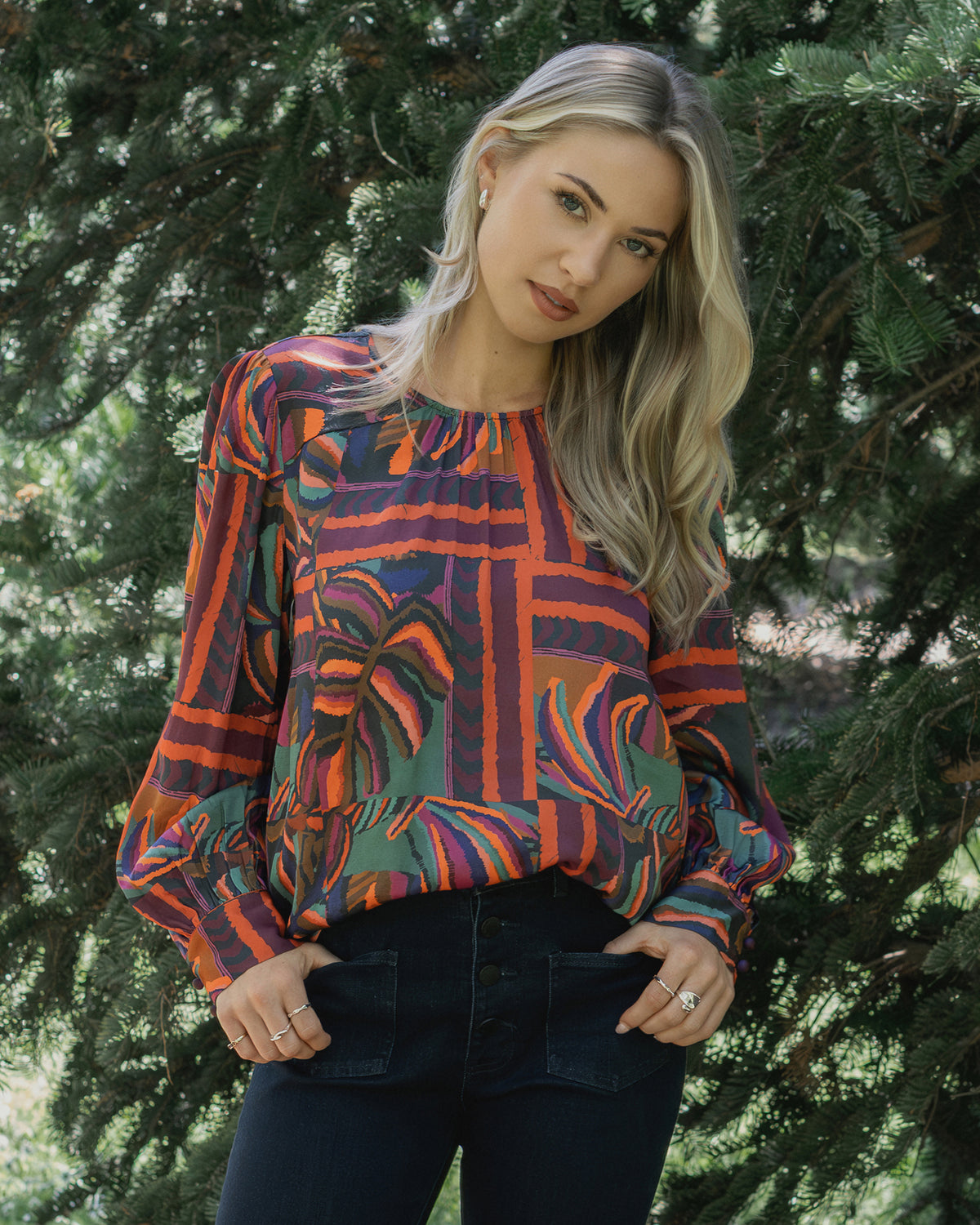 A woman with long blonde hair stands outdoors by green pine trees, wearing the Knewe Label Lexie Top in Sunset, featuring hues of red, purple, and orange, paired with dark pants. She looks relaxed with her hands in her pockets.