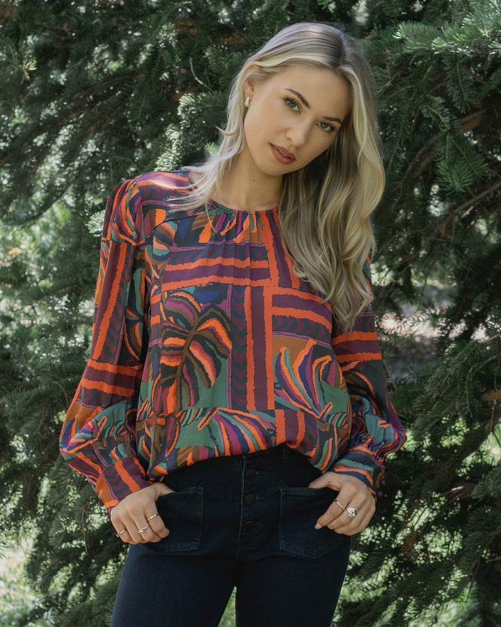 A woman with long blonde hair stands outdoors by green pine trees, wearing the Knewe Label Lexie Top in Sunset, featuring hues of red, purple, and orange, paired with dark pants. She looks relaxed with her hands in her pockets.