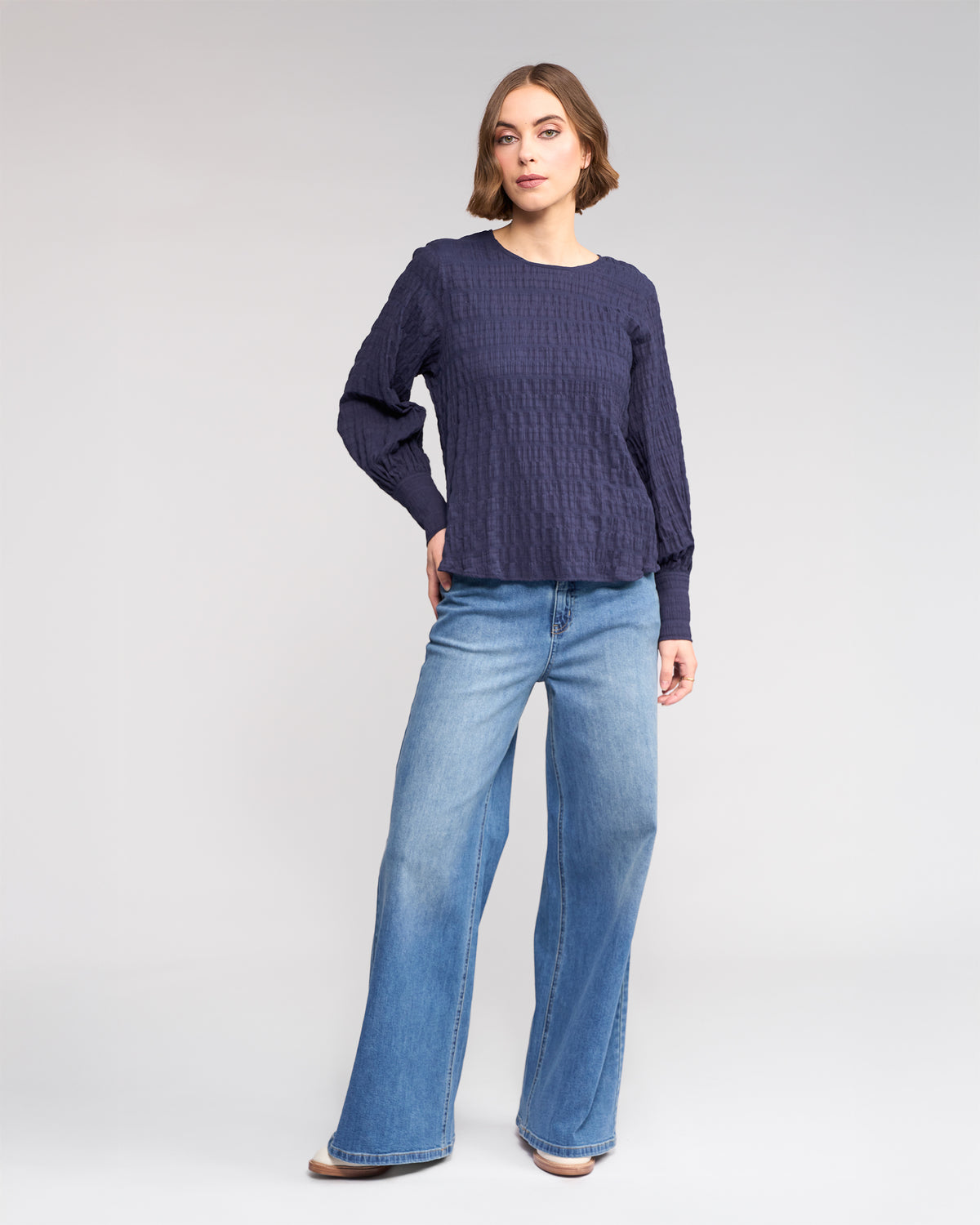 A woman poses against a plain background, wearing the Knewe Label Volume Top in Ink—a dark blue, long sleeve, textured cotton top—paired with light blue wide-leg jeans and tan shoes.