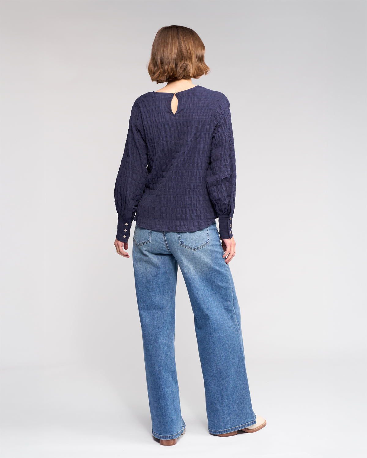 A person with short brown hair, wearing the Knewe Label Volume Top in Ink and wide-leg blue jeans, stands facing away against a plain light gray background.