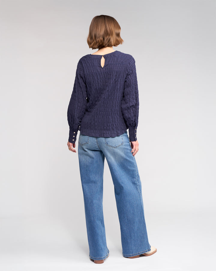 A person with short brown hair, wearing the Knewe Label Volume Top in Ink and wide-leg blue jeans, stands facing away against a plain light gray background.