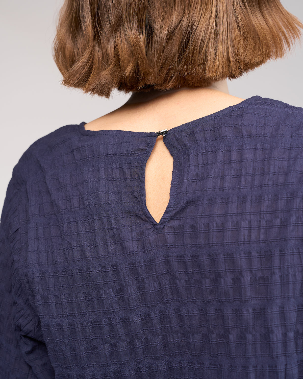 A close-up of a person with short brown hair wearing the Knewe Label Volume Top - Ink, featuring stretchy textured cotton, a small keyhole opening, and a button at the neckline.