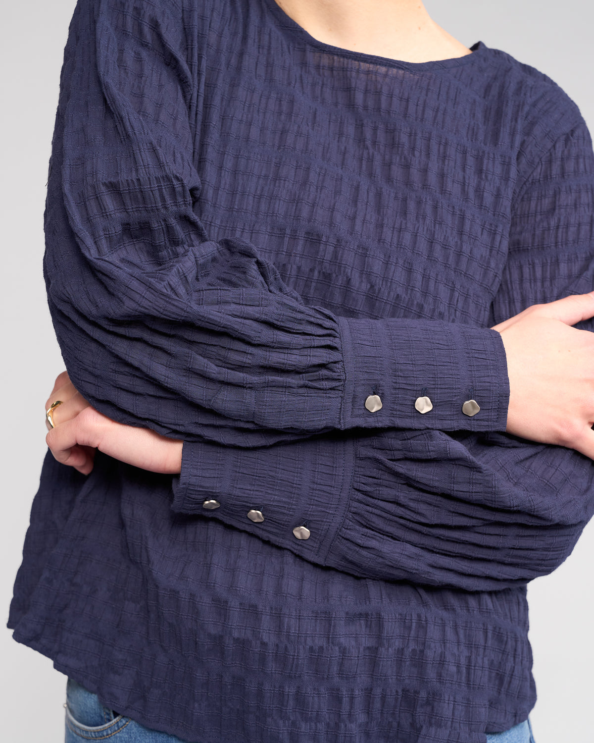 A person wearing the Knewe Label Volume Top in Ink, a dark blue textured long sleeve with pleated sleeves and metallic cuff buttons, stands with arms crossed. The head is partially out of frame.