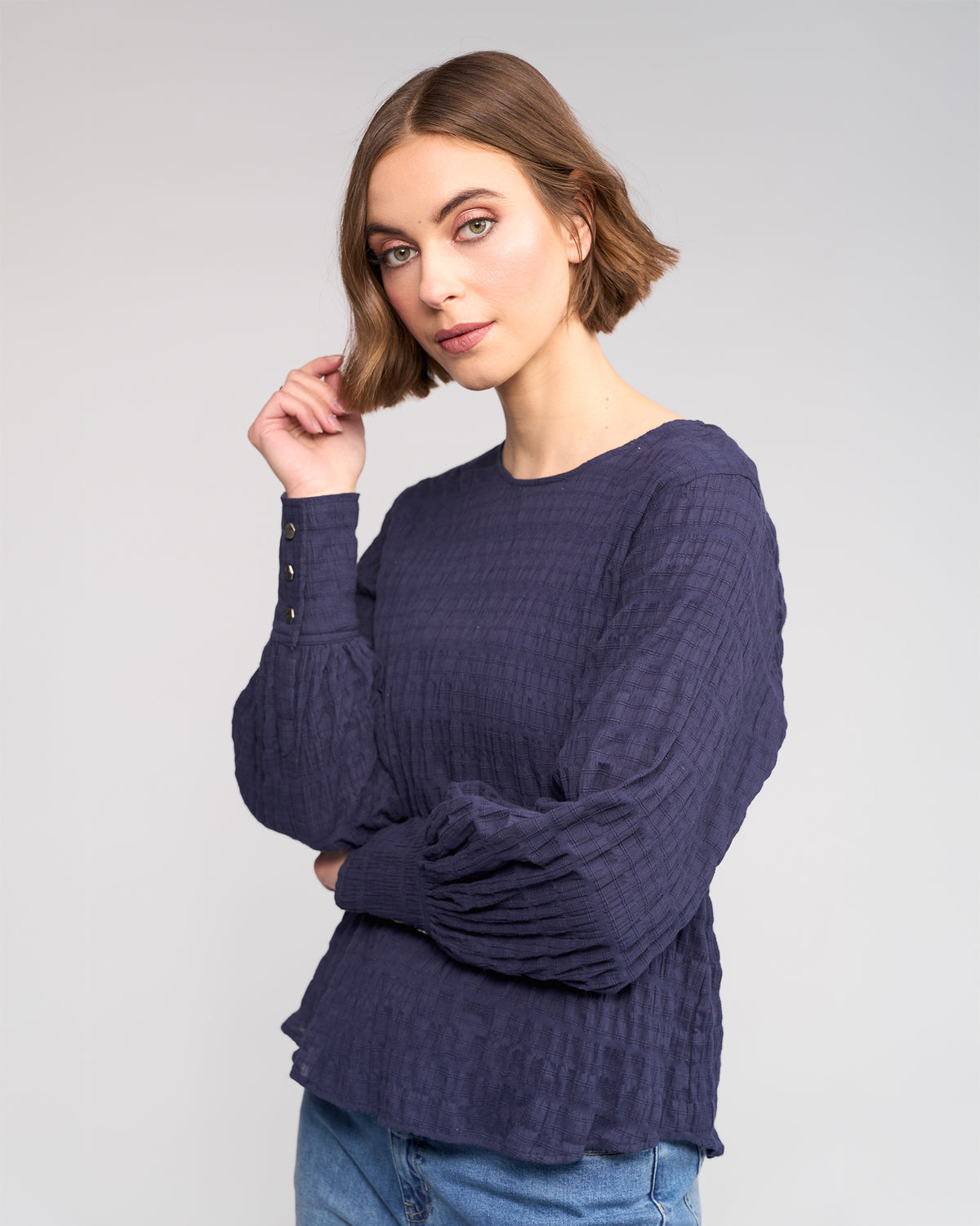 A woman with a short brown bob wears the Knewe Label Volume Top - Ink, a stretchy textured cotton long sleeve, paired with blue jeans. She stands against a plain light background, one hand touching her hair.