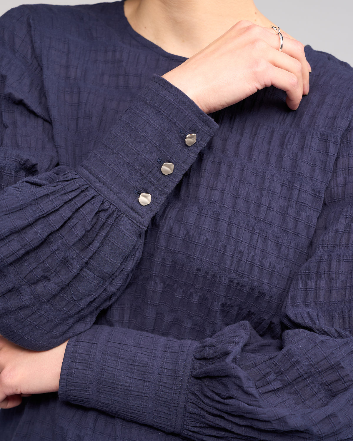 A person wearing the Knewe Label Volume Top in Ink, featuring pleated long sleeves, three metallic cuff buttons, and textured fabric, rests one hand on their shoulder.