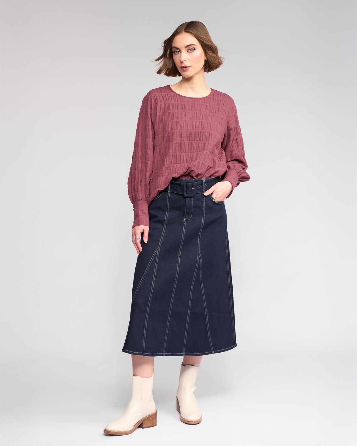 A woman with short brown hair and a neutral expression wears the Knewe Label Volume Top in Port, featuring stretchy textured cotton and a round neckline. She pairs it with a high-waisted dark denim midi skirt and beige ankle boots.
