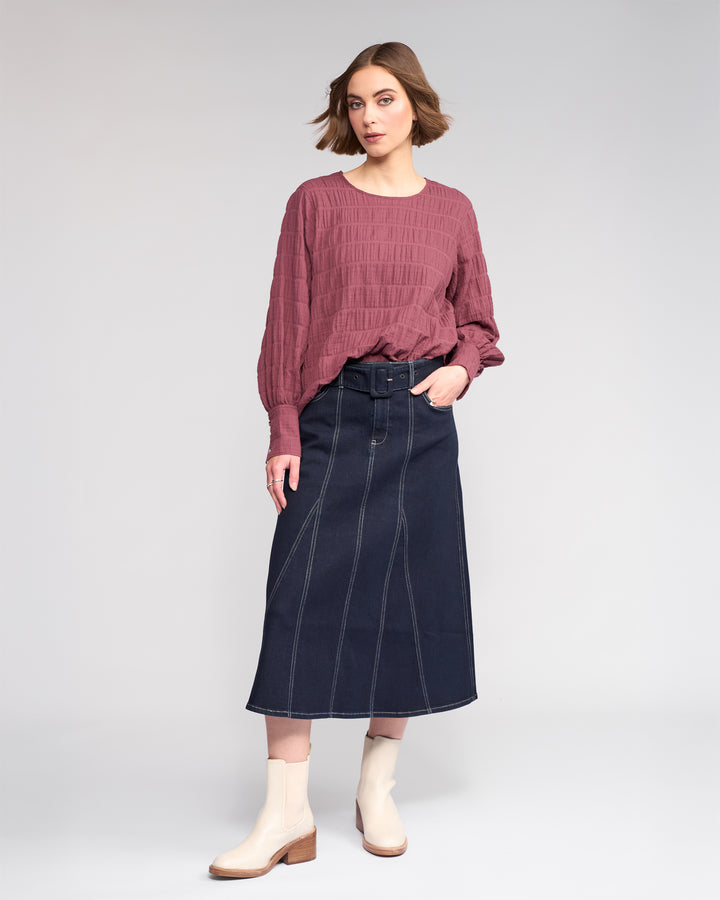 A woman with short brown hair and a neutral expression wears the Knewe Label Volume Top in Port, featuring stretchy textured cotton and a round neckline. She pairs it with a high-waisted dark denim midi skirt and beige ankle boots.
