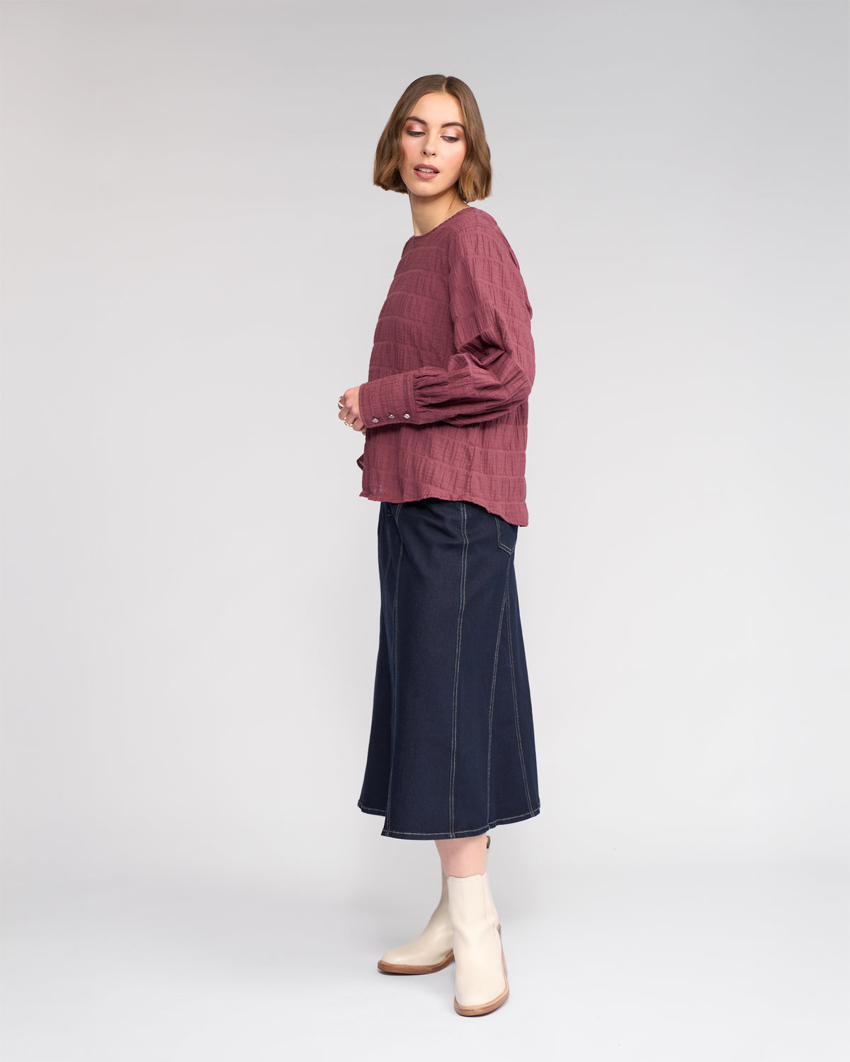 A woman stands sideways against a plain light gray background, wearing the Knewe Label Volume Top - Port, paired with a dark denim midi skirt and light ankle boots.