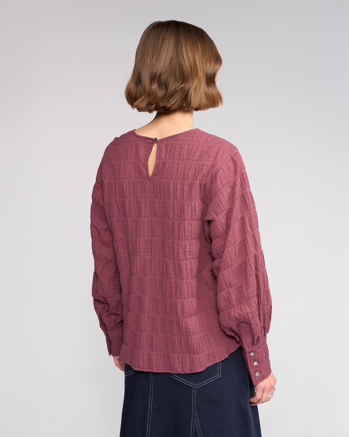 A person with short brown hair, seen from behind, wears the Knewe Label Volume Top - Port with a keyhole opening at the round neckline and buttoned cuffs, paired with a dark skirt against a plain light gray background.