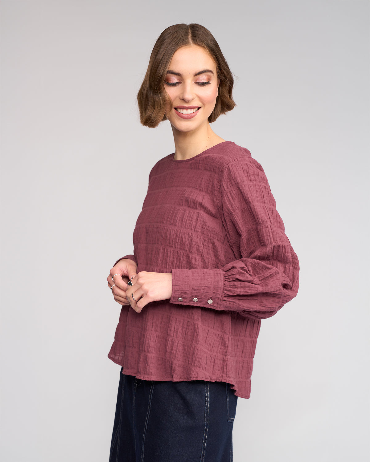 A woman with short brown hair wears the Knewe Label Volume Top in Port, a stretchy textured cotton long-sleeve, paired with dark pants. She stands sideways, smiling softly and looking down against a plain light gray background.