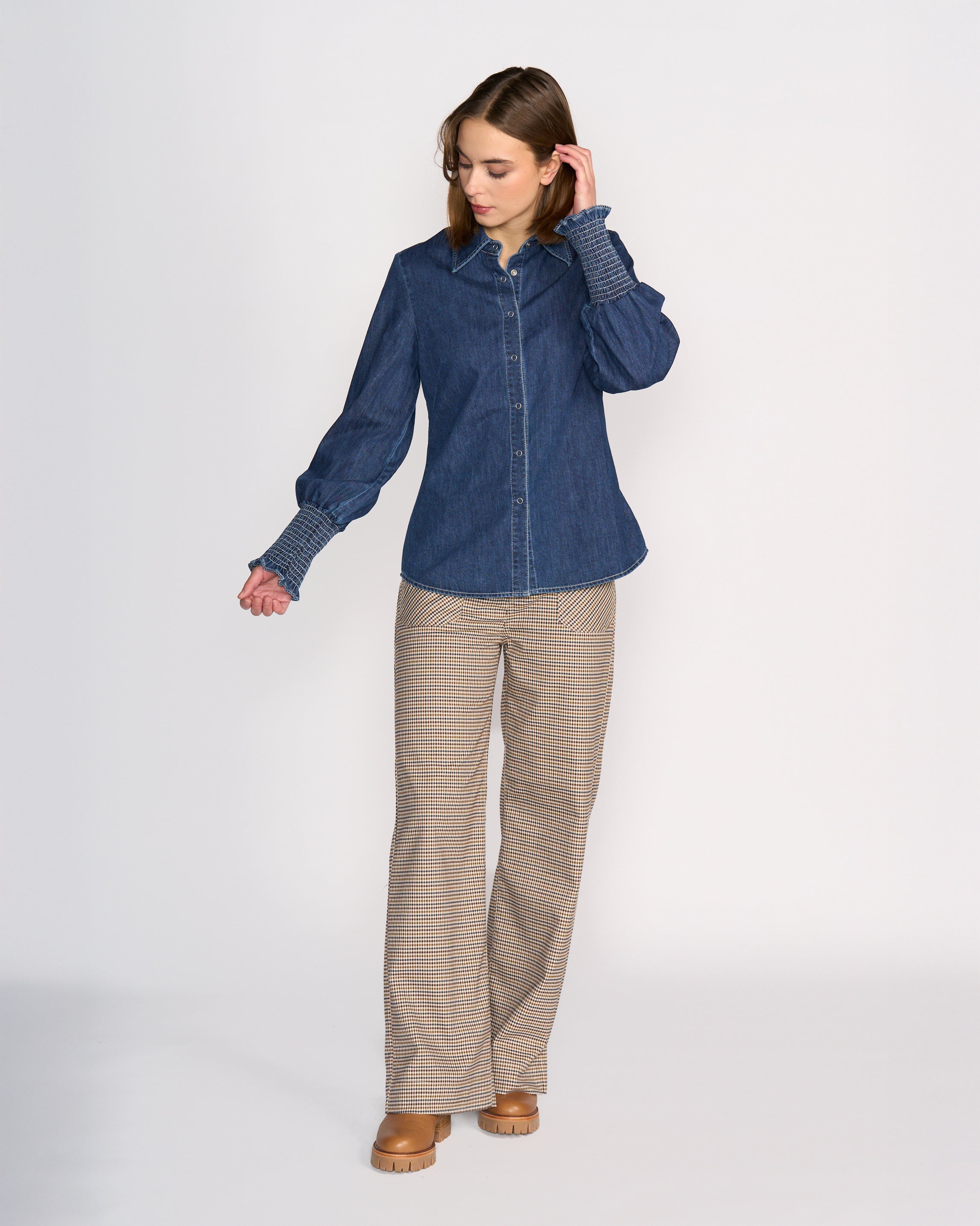 A woman stands against a plain background wearing Knewe Label’s Ford Shirt in dark blue 100% cotton denim, paired with plaid wide-leg pants and tan platform shoes, looking down with one hand in her hair.