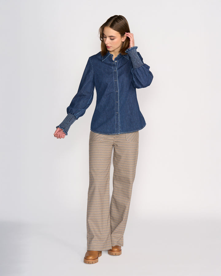 A woman stands against a plain background wearing Knewe Label’s Ford Shirt in dark blue 100% cotton denim, paired with plaid wide-leg pants and tan platform shoes, looking down with one hand in her hair.