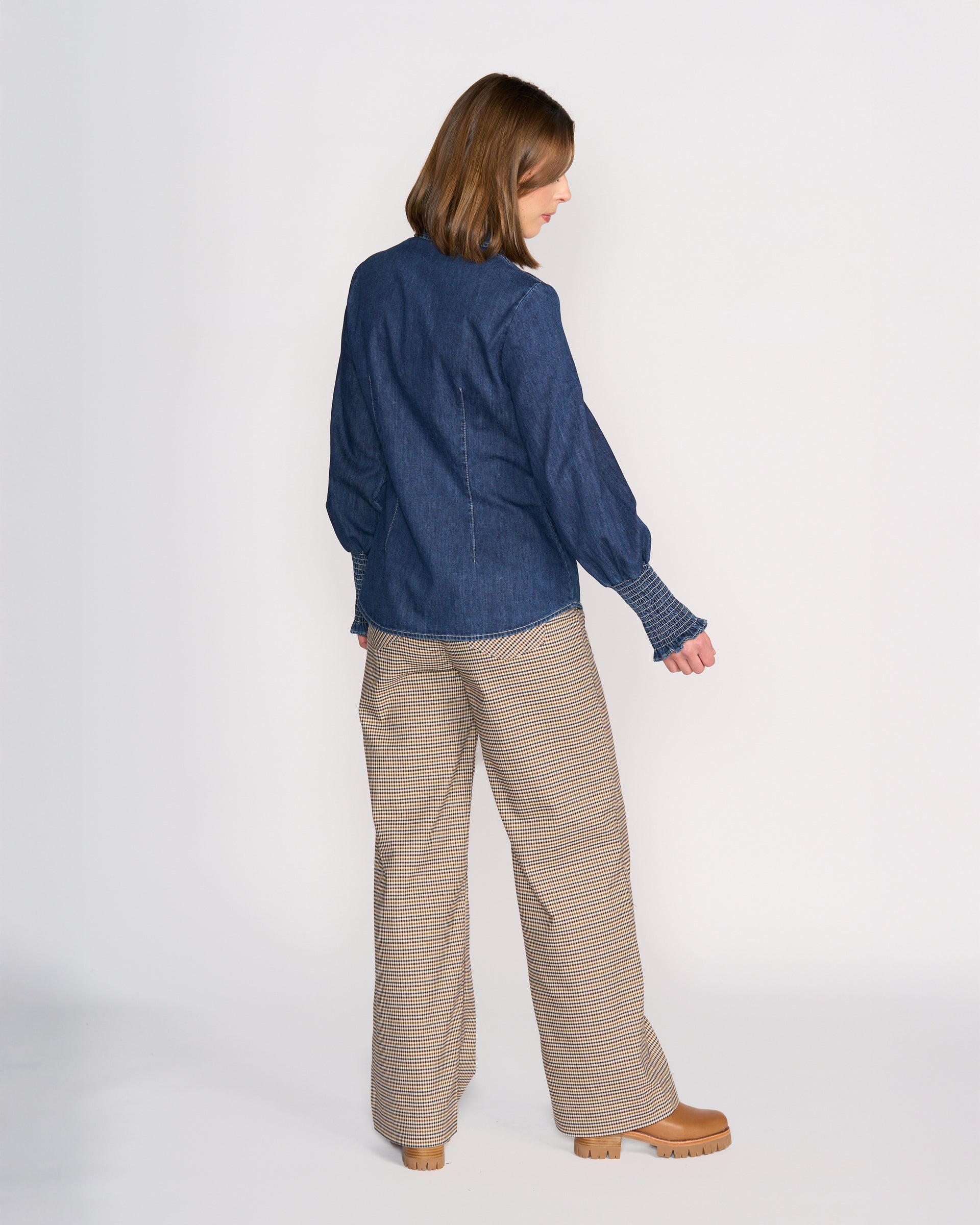 A woman faces away from the camera wearing the Knewe Label Ford Shirt in dark blue and wide-leg plaid pants with tan platform shoes, set against a plain white background.