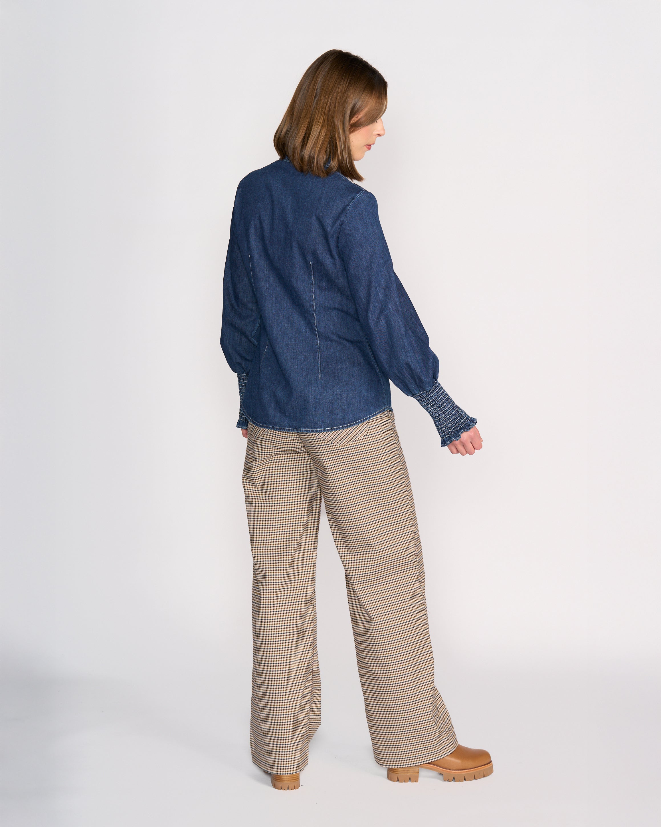 A woman faces away from the camera wearing the Knewe Label Ford Shirt in dark blue and wide-leg plaid pants with tan platform shoes, set against a plain white background.