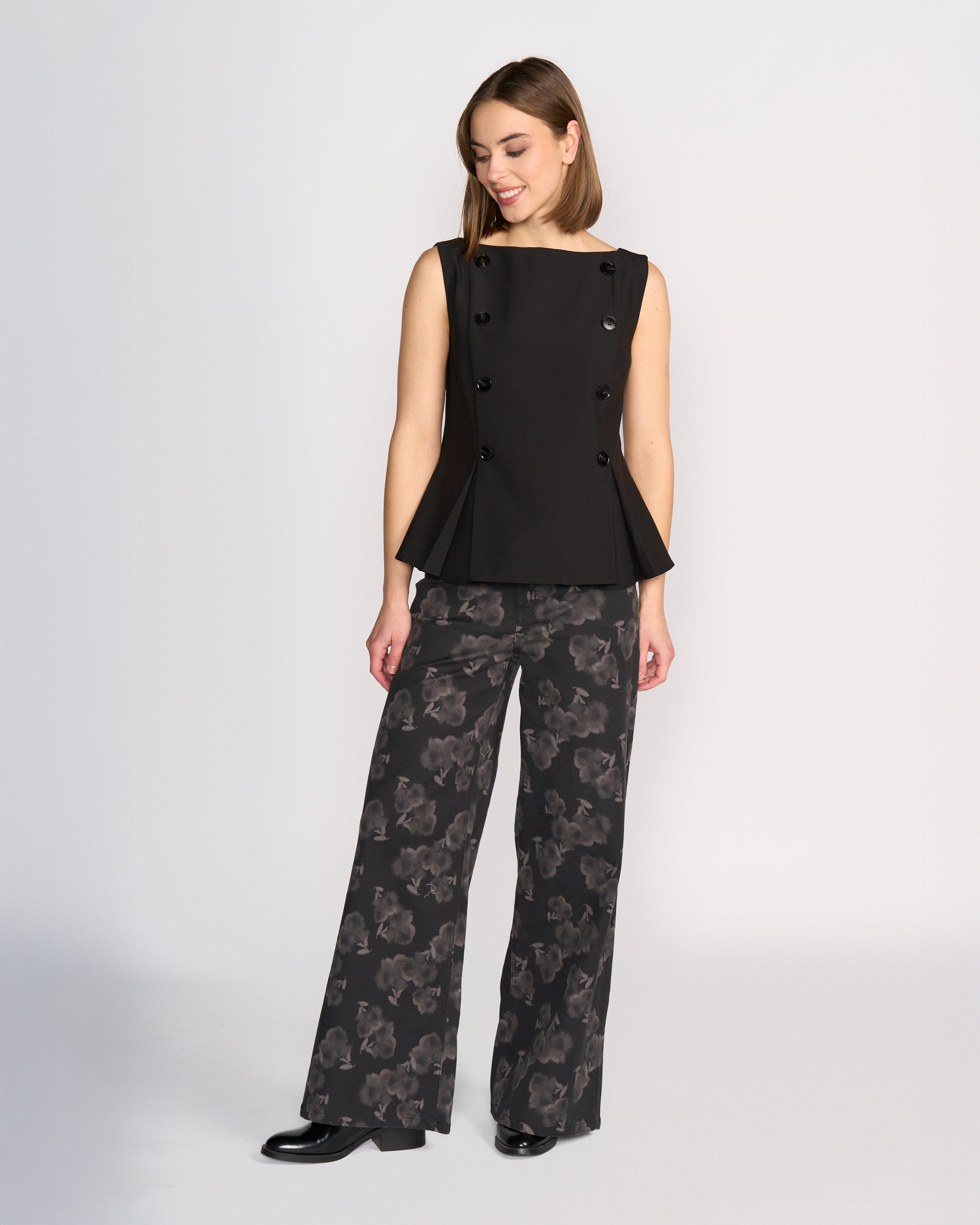 A woman stands against a plain light background wearing the Knewe Label Austin Top in black, a sleeveless vest with black button detailing, paired with wide-leg dark floral print pants. She looks down and smiles, arms relaxed at her sides.