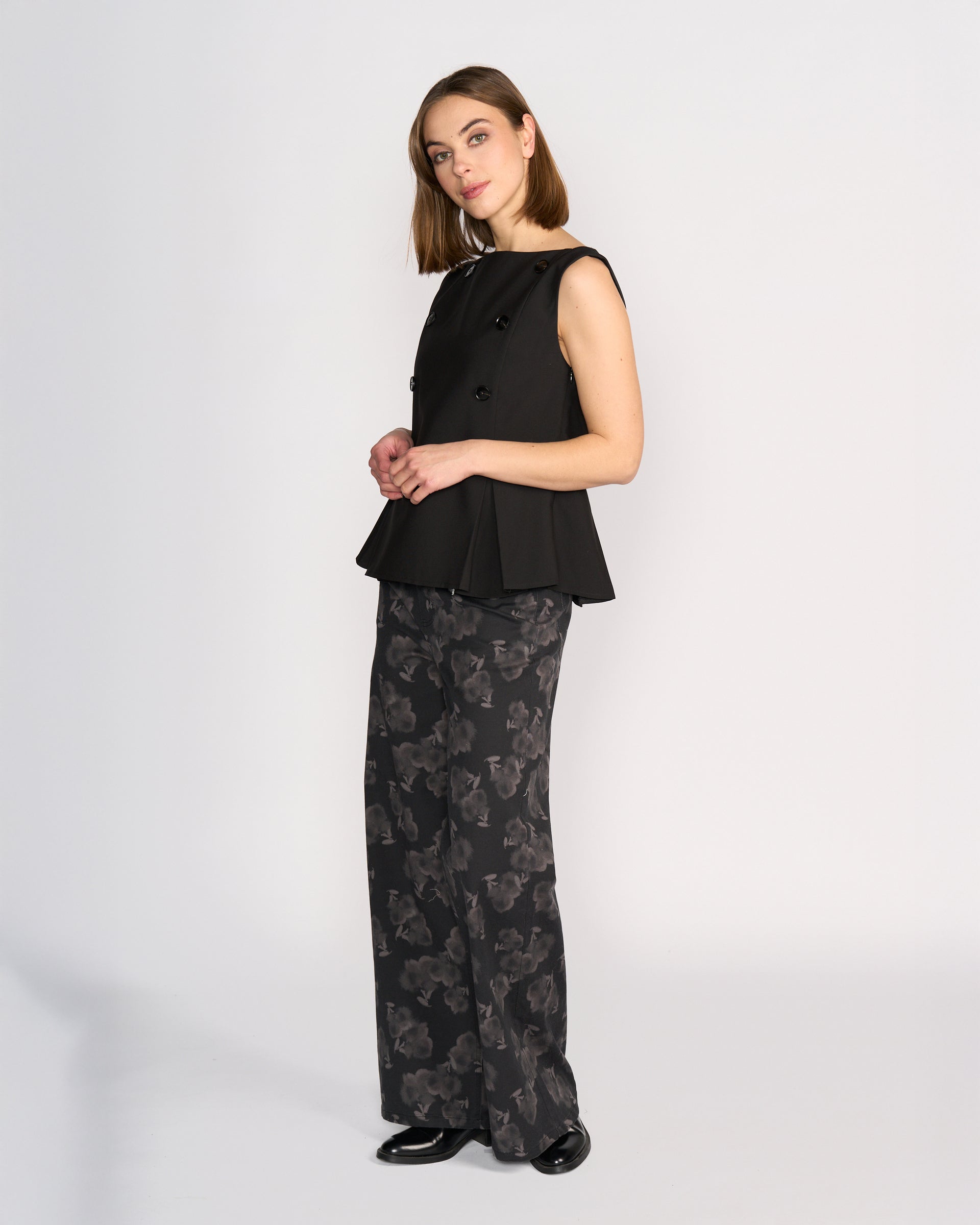 A woman wears the Knewe Label Austin Top in black, a sleeveless vest with pleat details and buttons, styled with wide-leg black pants featuring a gray abstract floral print and black shoes, standing against a plain white background.