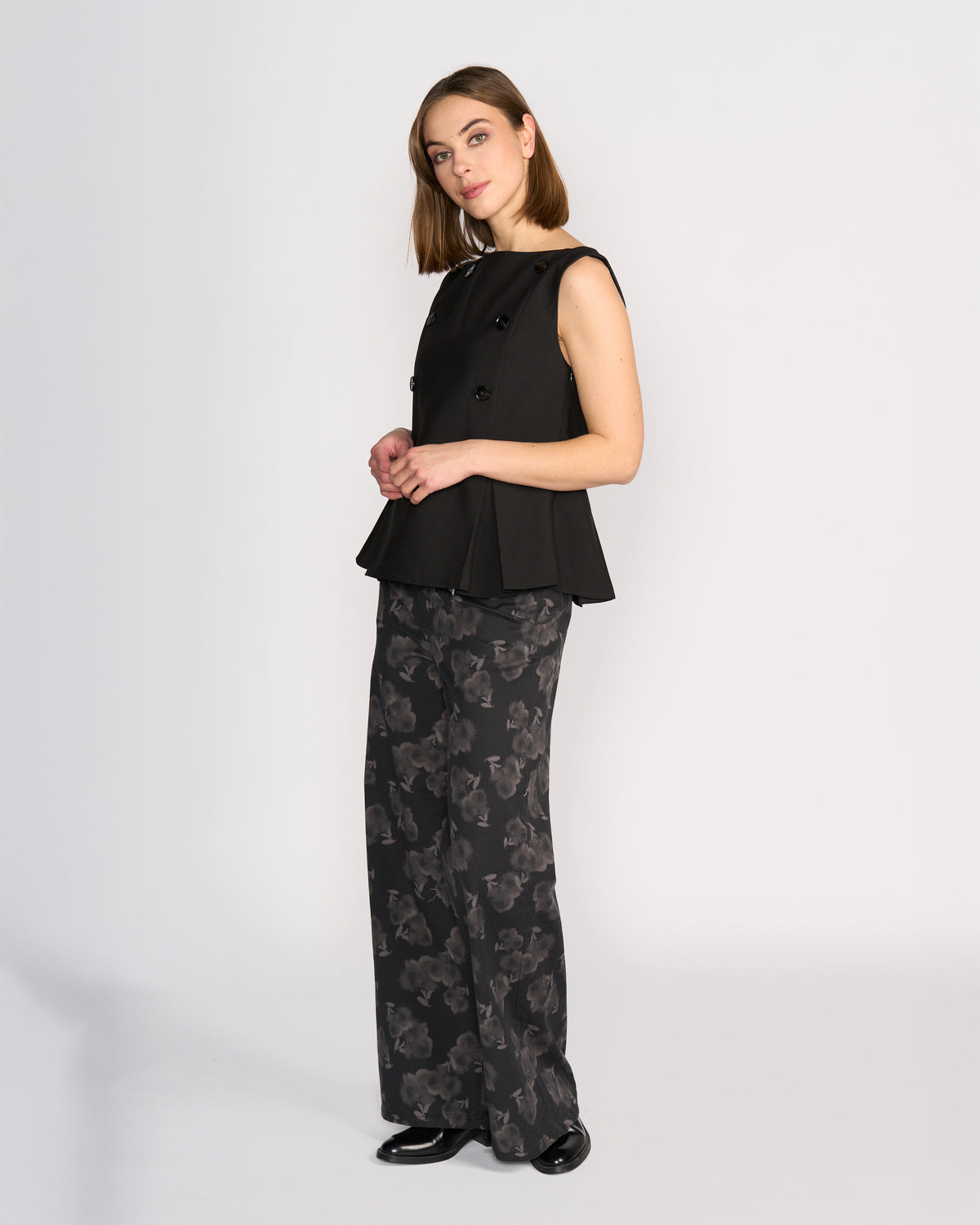 A woman wears the Knewe Label Austin Top in black, a sleeveless vest with pleat details and buttons, styled with wide-leg black pants featuring a gray abstract floral print and black shoes, standing against a plain white background.
