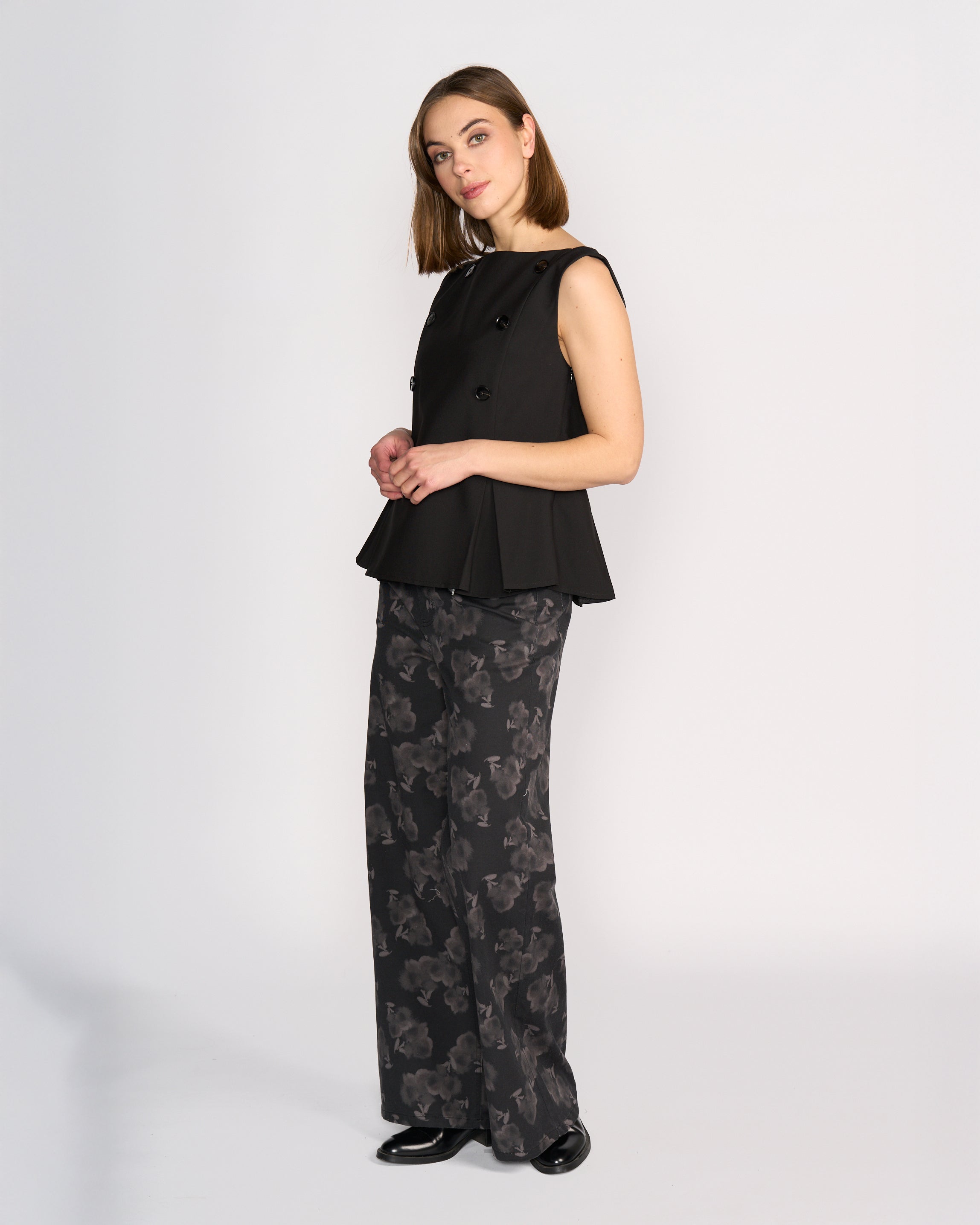 A woman wears the Knewe Label Austin Top in black, a sleeveless vest with pleat details and buttons, styled with wide-leg black pants featuring a gray abstract floral print and black shoes, standing against a plain white background.
