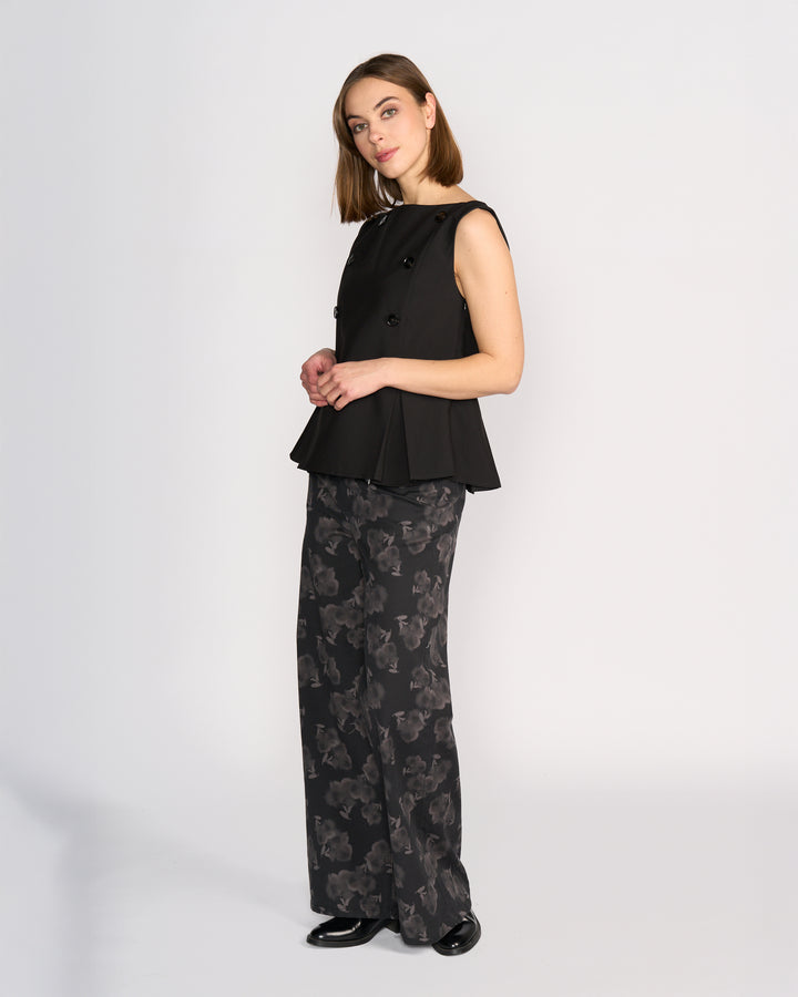 A woman wears the Knewe Label Austin Top in black, a sleeveless vest with pleat details and buttons, styled with wide-leg black pants featuring a gray abstract floral print and black shoes, standing against a plain white background.