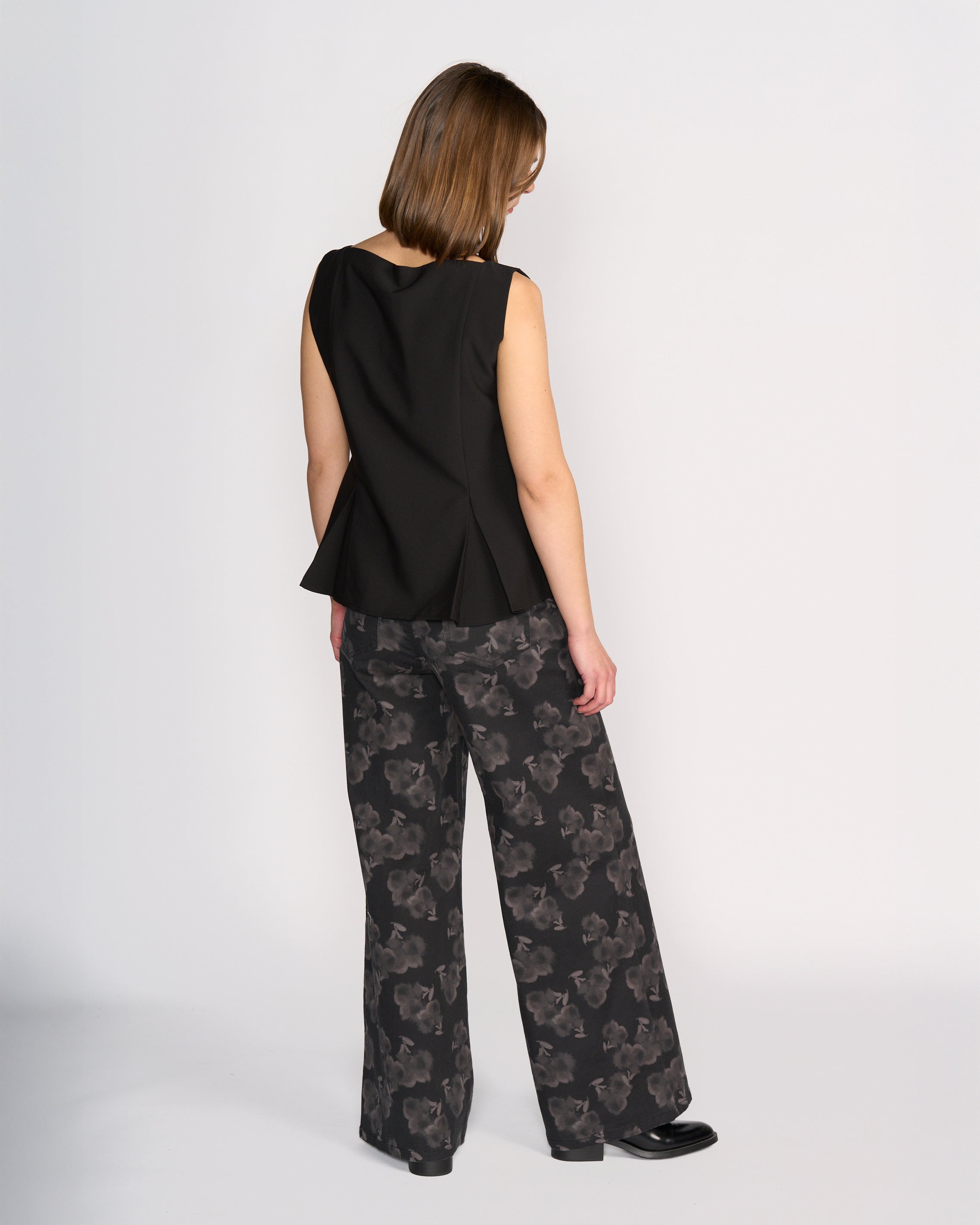 A woman faces away, wearing the Knewe Label Austin Top in black—a sleeveless vest with black button details and a flared hem—paired with wide-leg black pants featuring a gray floral pattern, set against a plain light background.