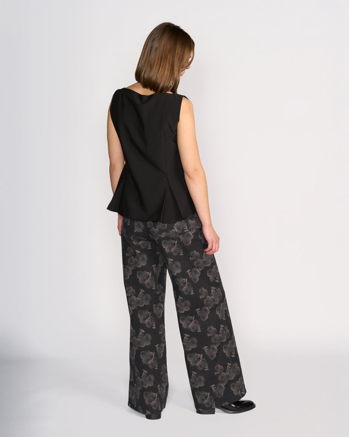 A woman faces away, wearing the Knewe Label Austin Top in black—a sleeveless vest with black button details and a flared hem—paired with wide-leg black pants featuring a gray floral pattern, set against a plain light background.