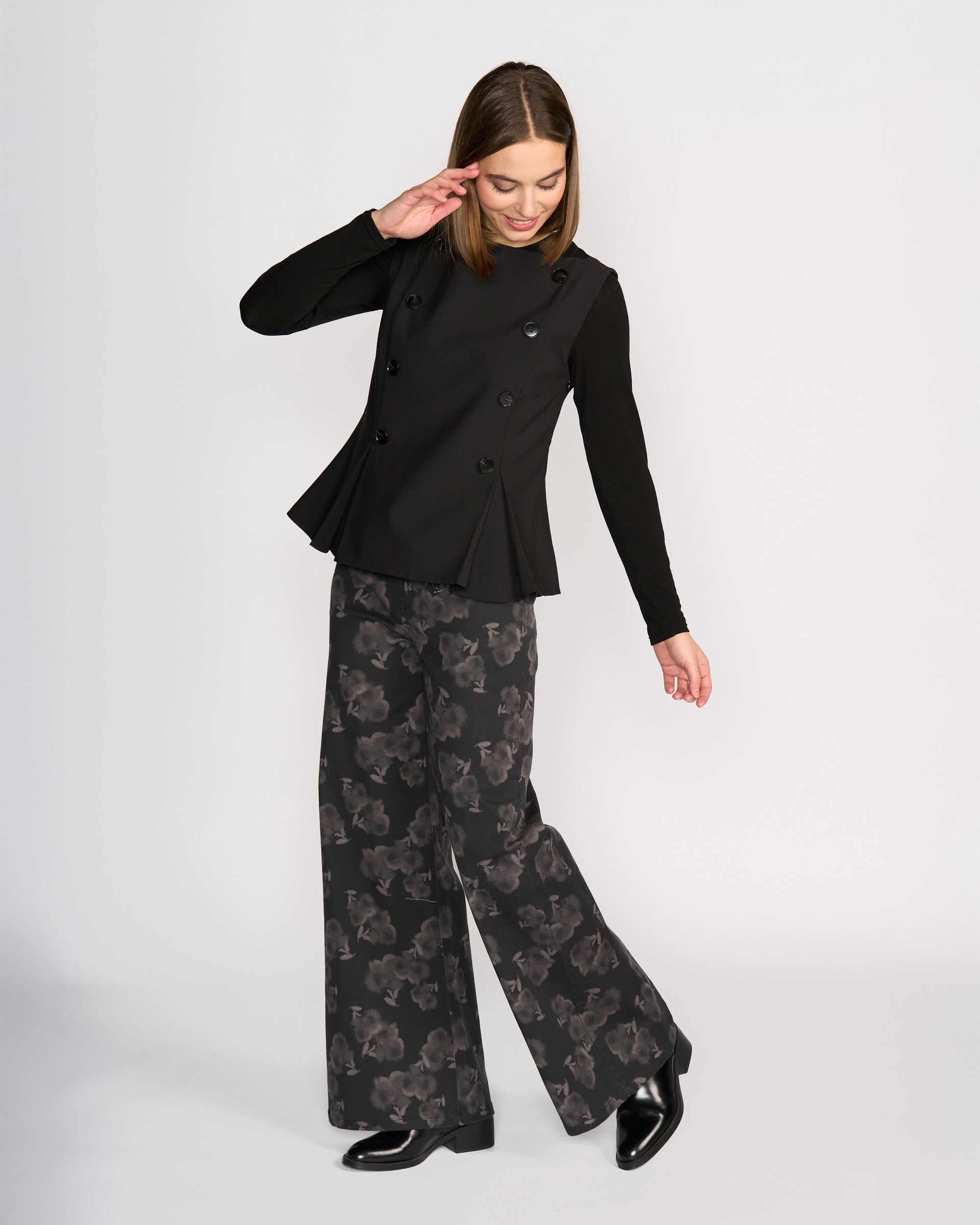 A woman smiles and tilts her head, raising one arm as she models the Knewe Label Austin Top in black against a plain background, paired with wide-leg black pants featuring a subtle floral pattern.