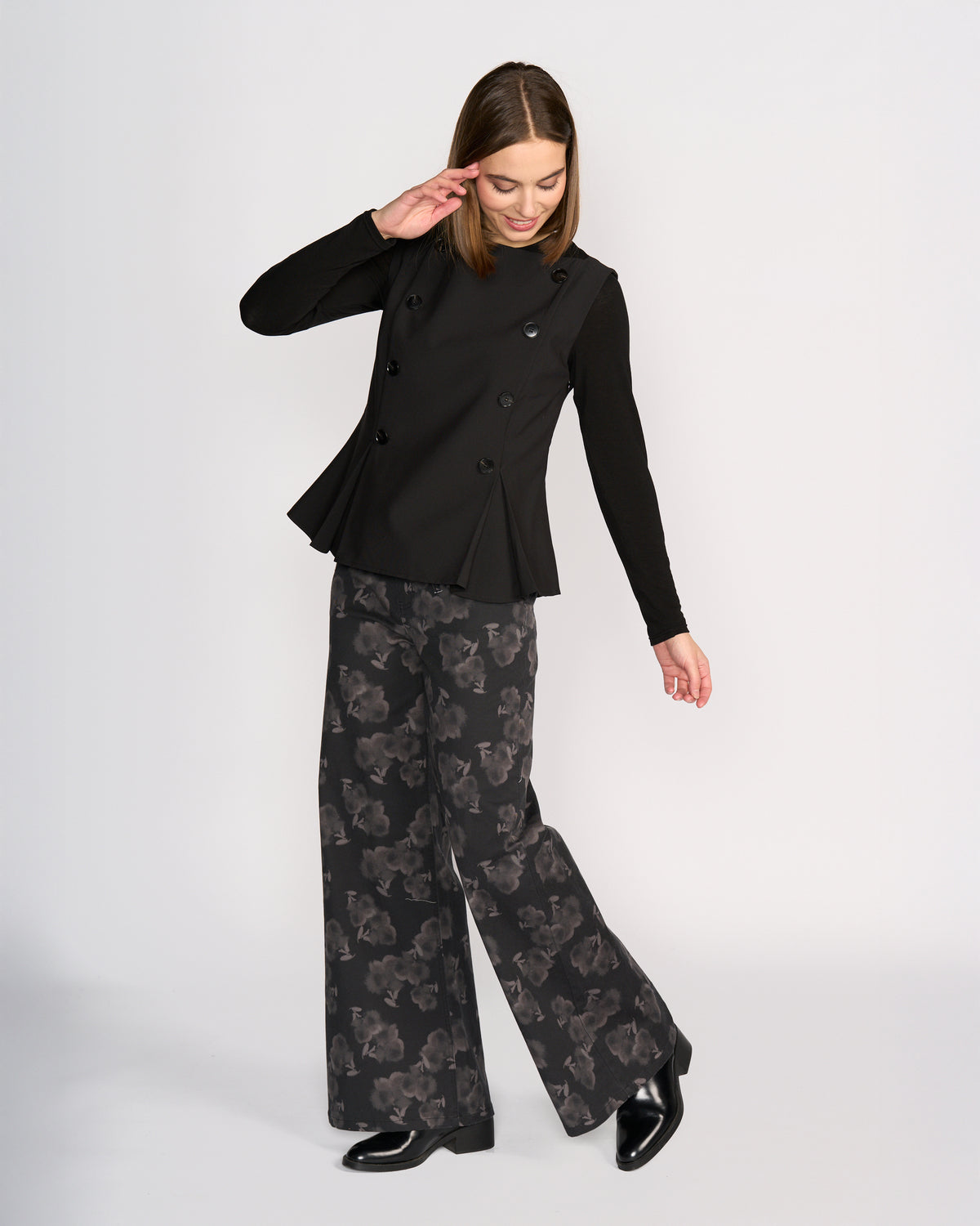 A woman smiles and tilts her head, raising one arm as she models the Knewe Label Austin Top in black against a plain background, paired with wide-leg black pants featuring a subtle floral pattern.