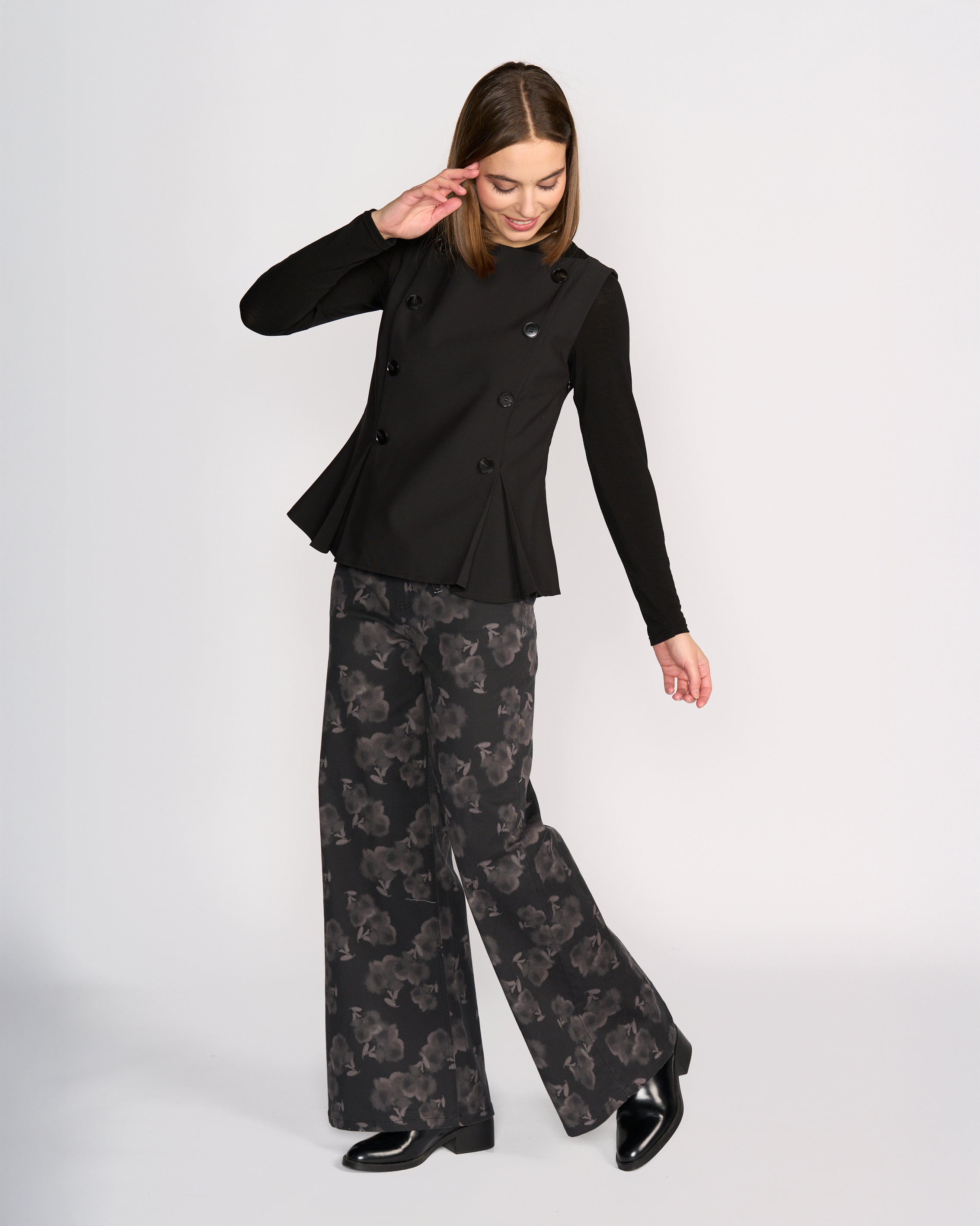A woman smiles and tilts her head, raising one arm as she models the Knewe Label Austin Top in black against a plain background, paired with wide-leg black pants featuring a subtle floral pattern.