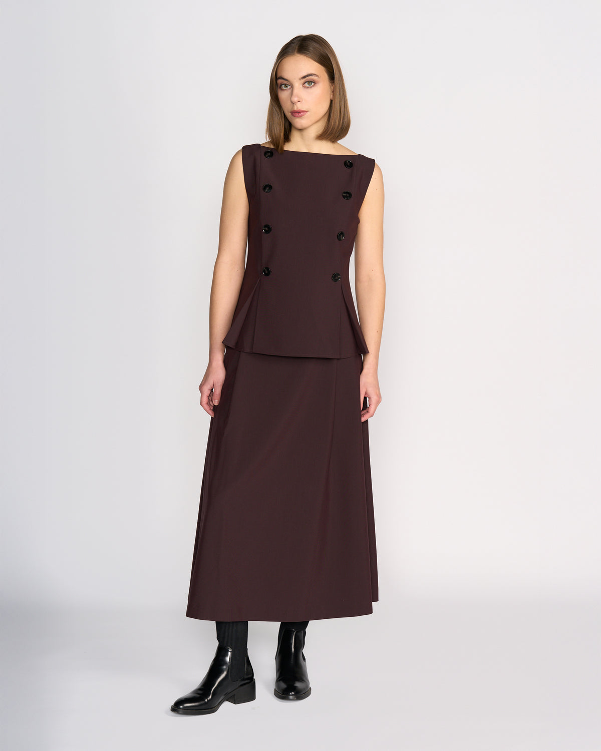 A woman stands in a studio wearing the Knewe Label Austin Top in Raisin, a sleeveless vest top with black buttons and pleat details, paired with a matching long skirt, black ankle boots, and straight shoulder-length hair for a tailored look.