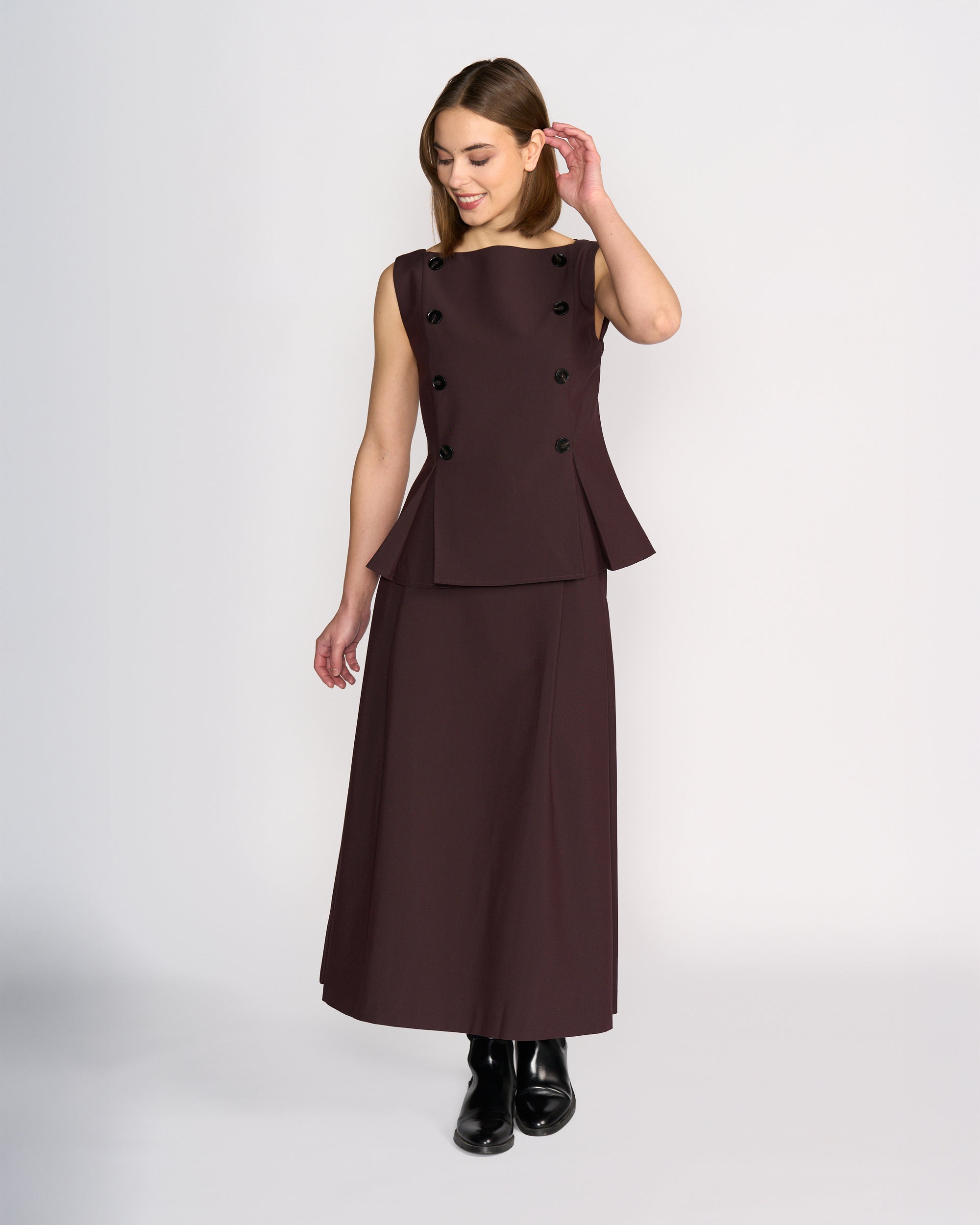 A woman wears the Knewe Label Austin Top in Raisin, a sleeveless tailored vest with pleat details, paired with a matching long skirt. She looks down, touches her hair, and completes the look with black ankle boots.