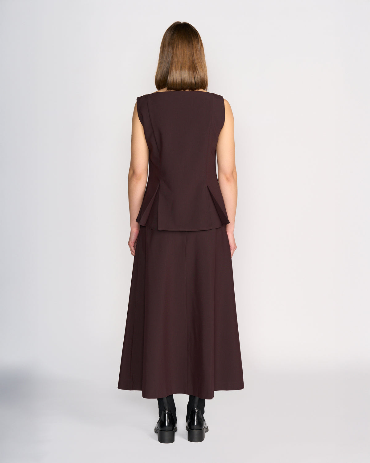A person with straight, shoulder-length brown hair is seen from behind wearing the Knewe Label Austin Top in Raisin, featuring subtle peplum and pleat details, paired with a matching long skirt and black boots.