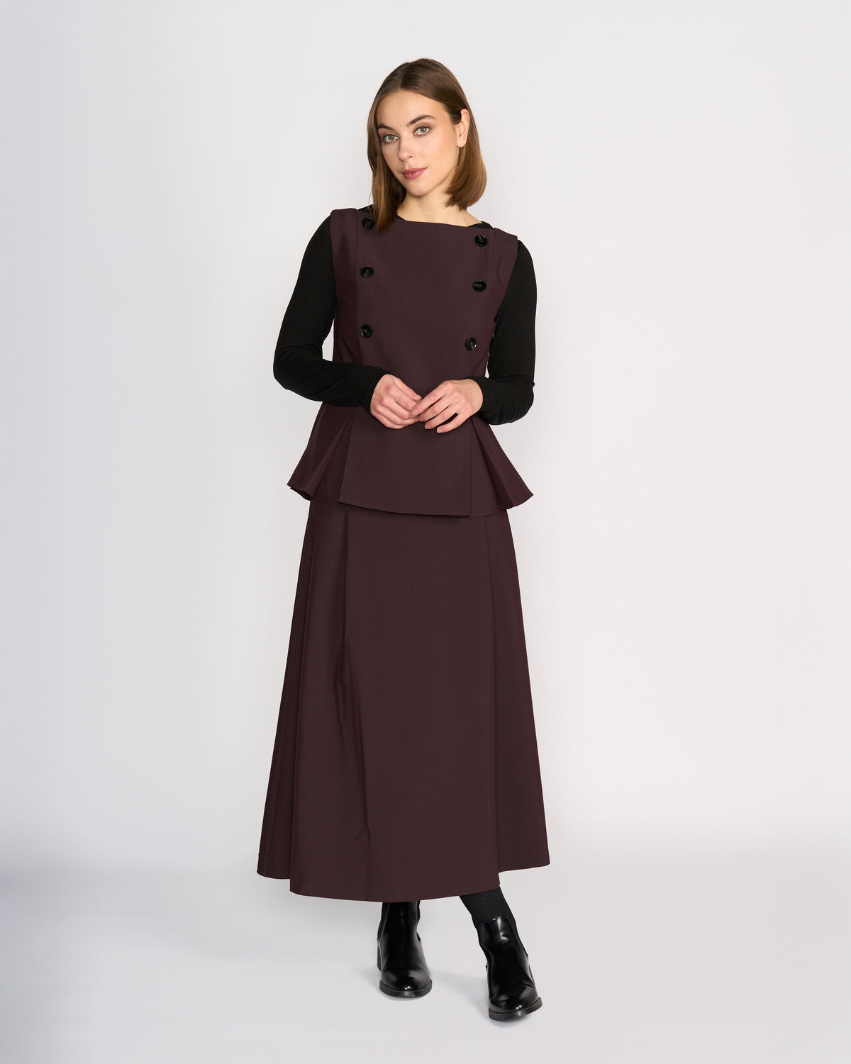A woman stands against a plain light background wearing the Knewe Label Austin Top in Raisin over a long-sleeve black top, paired with black ankle boots. She looks slightly to the side with a neutral expression.