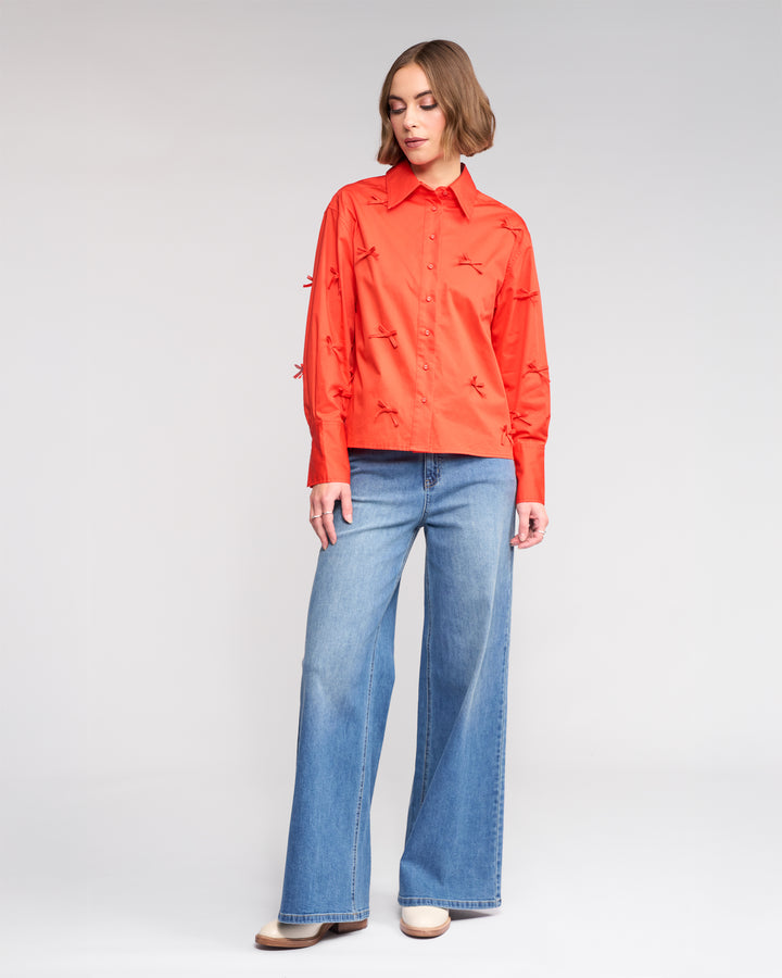 A woman with short brown hair models the Knewe Label Bow Shirt - Lava, a bright orange cotton top with embroidered details, paired with light blue wide-leg jeans and beige shoes, standing against a plain white backdrop.