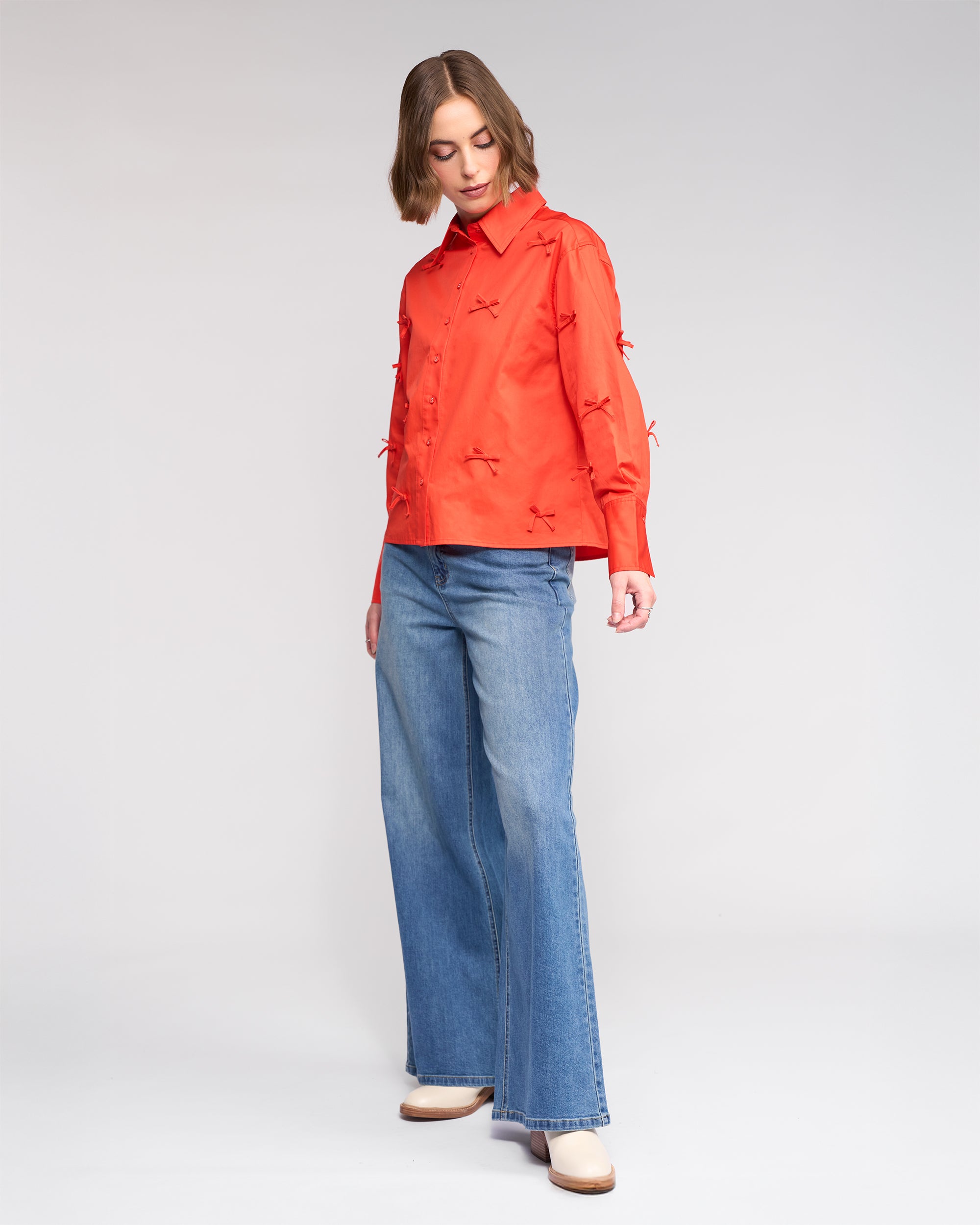 A woman wears the Knewe Label Bow Shirt - Lava with wide-leg blue jeans and beige shoes, standing against a plain light background. She looks down with one hand by her side.