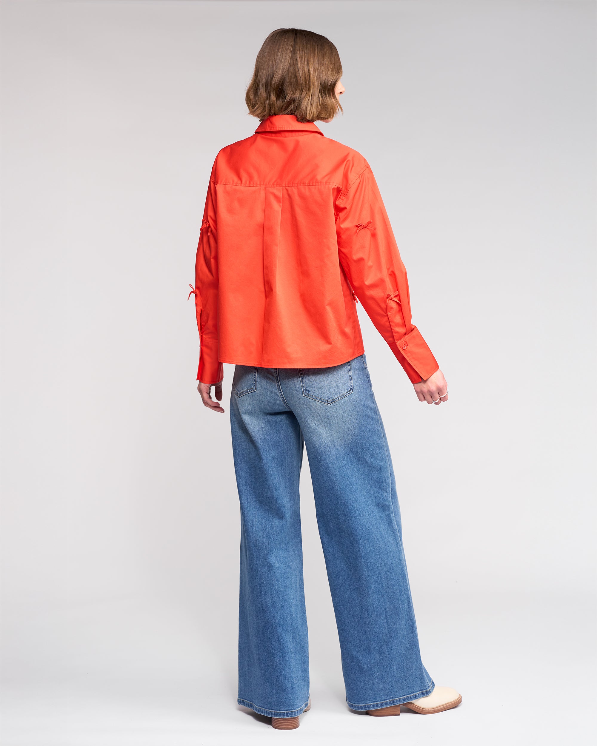 A person with chin-length brown hair faces away, wearing Knewe Label's Bow Shirt - Lava (bright orange), blue wide-leg jeans, and light-colored shoes against a plain light background.
