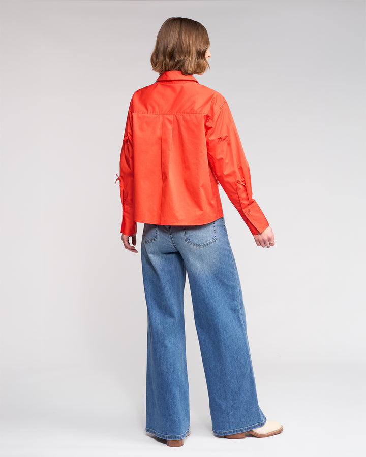 A person with chin-length brown hair faces away, wearing Knewe Label's Bow Shirt - Lava (bright orange), blue wide-leg jeans, and light-colored shoes against a plain light background.